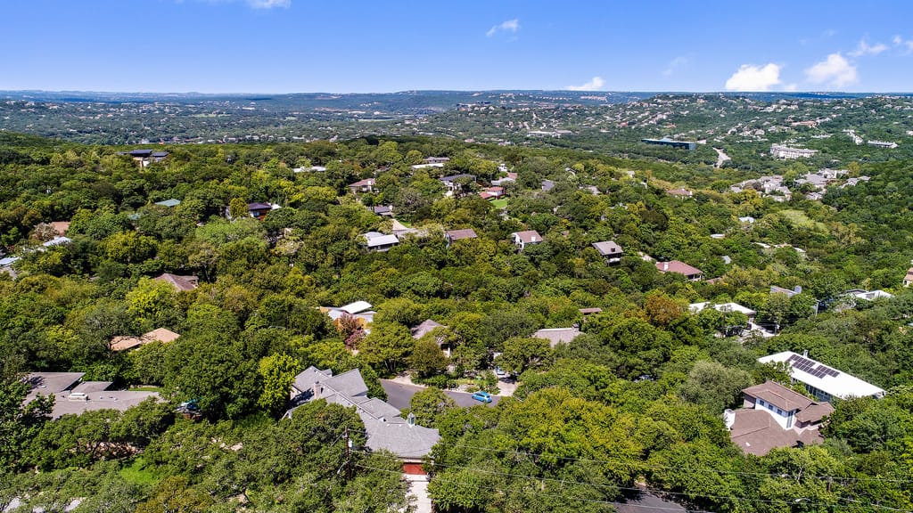 Aerial over Central Austin houses under heavy tree canopy, the Hill Country low on the horizon