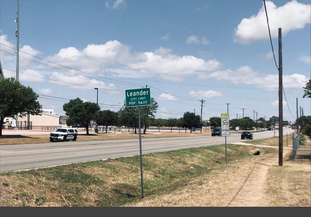 A Leander city limit sign beside a divided highway under a wide Texas sky