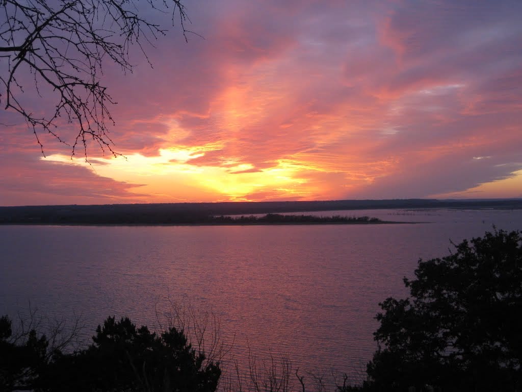 Sunset over open water at Lake Whitney, a low shoreline and the sun breaking under a band of cloud