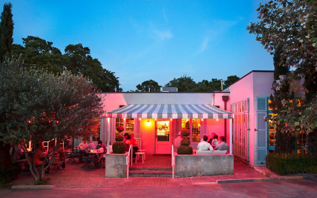 A pink-walled cafe at dusk in Bouldin Creek, a striped awning over a patio of diners under an olive tree