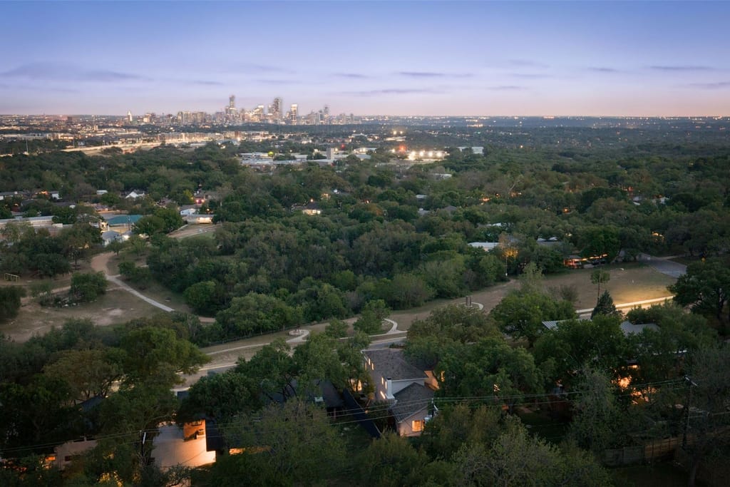 Aerial view at dusk over wooded streets and a creekside park, with the lit downtown Austin skyline beyond