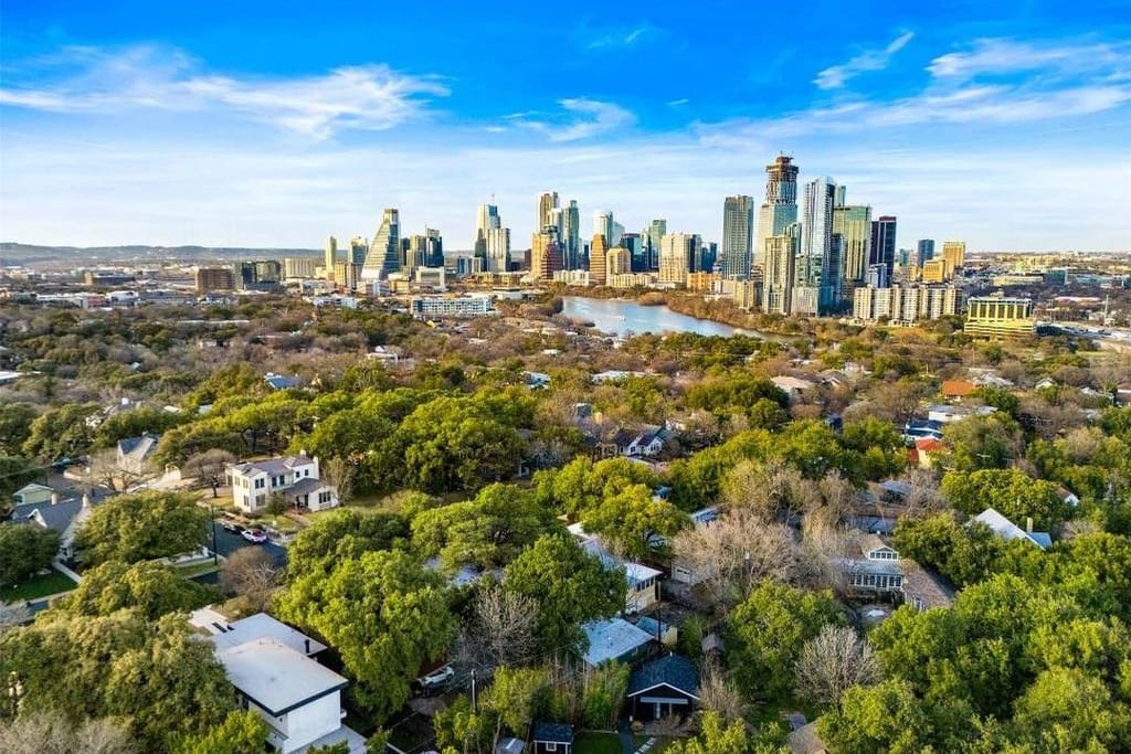 Aerial view over tree-lined streets and houses toward Lady Bird Lake and the downtown Austin skyline
