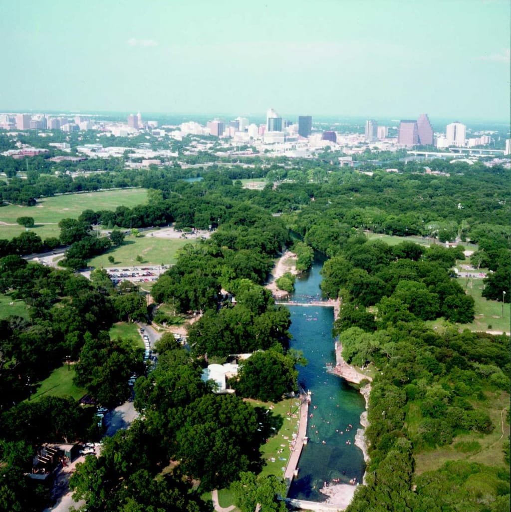 Barton Springs Pool from above, the spring fed channel green between its walls, the Austin skyline behind the trees